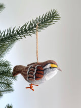 A charming White Throat Sparrow ornament, handcrafted from wool felt, hangs from a lush green Christmas tree branch. The unique Christmas ornament features a rich brown back, distinctive black and white crown stripes, a white throat, and bright yellow lores. Playful orange feet dangle below, with a jute string for easy hanging.