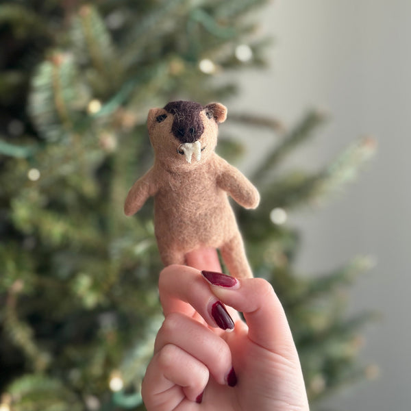 A woman's hand with dark red nail polish holds a handcrafted needle felted wombat finger puppet, displayed against a blurred green Christmas tree background. The unique wombat puppet features a stout, light brown body, small rounded ears, a dark brown head, and two distinct white teeth.