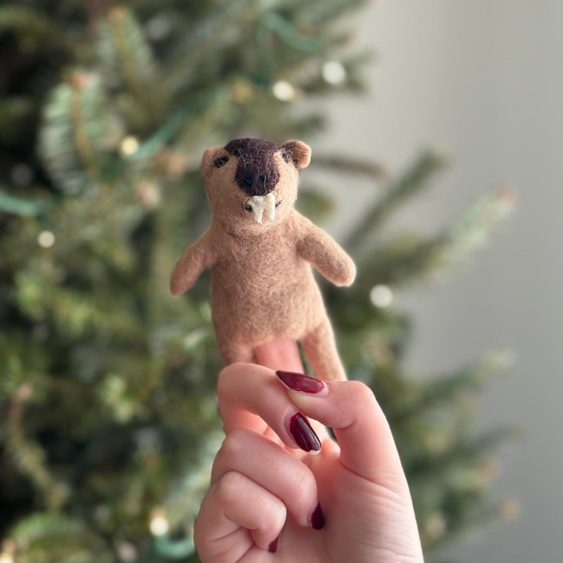 A woman's hand with dark red nail polish holds a handcrafted needle felted wombat finger puppet, displayed against a blurred green Christmas tree background. The unique wombat puppet features a stout, light brown body, small rounded ears, a dark brown head, and two distinct white teeth.