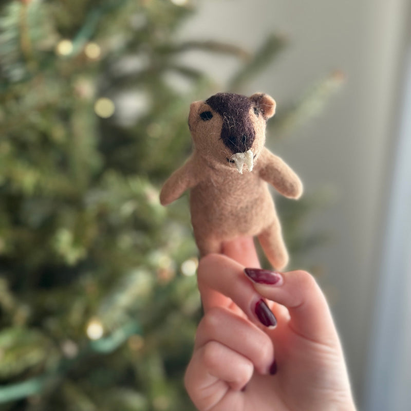A handcrafted needle felted wombat finger puppet is gently held by a hand, set against a blurred background of warm Christmas tree lights. This charming, compact 3D felt puppet is predominantly light brown with a dark brown head patch, tiny rounded ears, and small white teeth, highlighting its stout body. Its soft texture and unique design make it a delightful, festive toy.