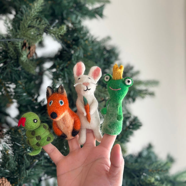 A hand presents a set of handmade felt woodland animal finger puppets against a festive Christmas tree. This unique collection, crafted by talented female artisans in Nepal, includes a green snake finger puppet with a red tongue, an orange and black fox finger puppet, a bunny finger puppet holding a felt carrot, and a frog finger puppet wearing a golden crown. These felt toys for kids bring whimsical joy.