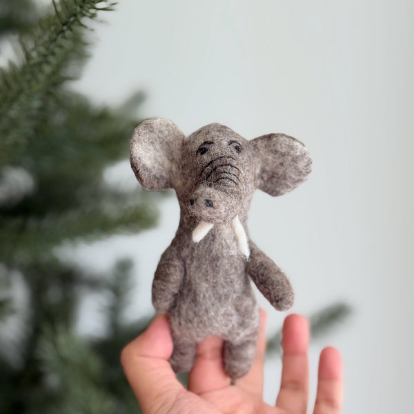 A handmade wool felt elephant finger puppet is held in a female hand against a blurred Christmas pine tree. This charming gray, needle felted elephant puppet features a detailed trunk, tusks, and ears, appearing soft and tactile.
