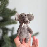 A handmade wool felt elephant finger puppet is held gently in a female hand, with a blurred Christmas tree in the background. This gray felt elephant features large ears, white tusks, and delicately stitched facial details. Its raised trunk highlights the unique design, and the visible wool felt texture emphasizes its handcrafted nature.