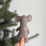 A light brown wool felt elephant finger puppet is worn on a female hand, set against a blurred Christmas tree and white wall. This charming handcrafted elephant finger puppet, made from 100% wool with 3D needle felting, features prominent ears, a rounded shape, and a distinct nose, showcasing its unique design and craftsmanship. It's an engaging felt toy for kids.