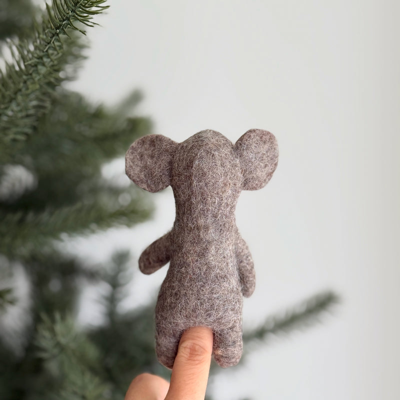 A light brown wool felt elephant finger puppet is worn on a female hand, set against a blurred Christmas tree and white wall. This charming handcrafted elephant finger puppet, made from 100% wool with 3D needle felting, features prominent ears, a rounded shape, and a distinct nose, showcasing its unique design and craftsmanship. It's an engaging felt toy for kids.