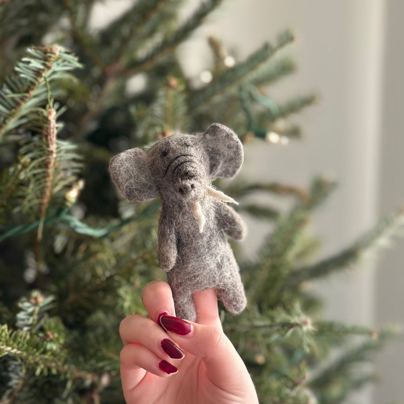 A light gray wool felt elephant finger puppet, handcrafted from textured wool, is held by a female hand with red polished nails. Its distinctive ears and tusks are visible against a blurred Christmas tree background adorned with lights, creating a cozy and festive scene.