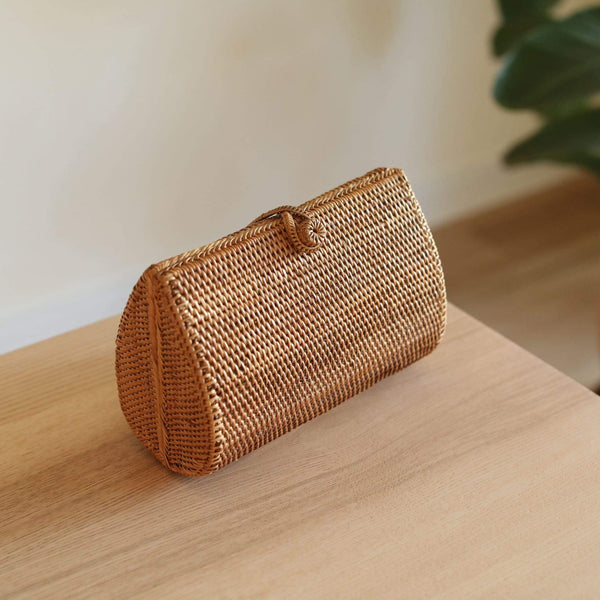A woven rattan clutch bag rests on a wooden surface, showcasing its natural beauty. The rectangular shape with rounded corners features an envelope-style closure with a hook, made from rattan or Ata grass with a textured weave texture. Inside, the lining displays a stunning batik print, adding to the bag's elegance and simplicity.