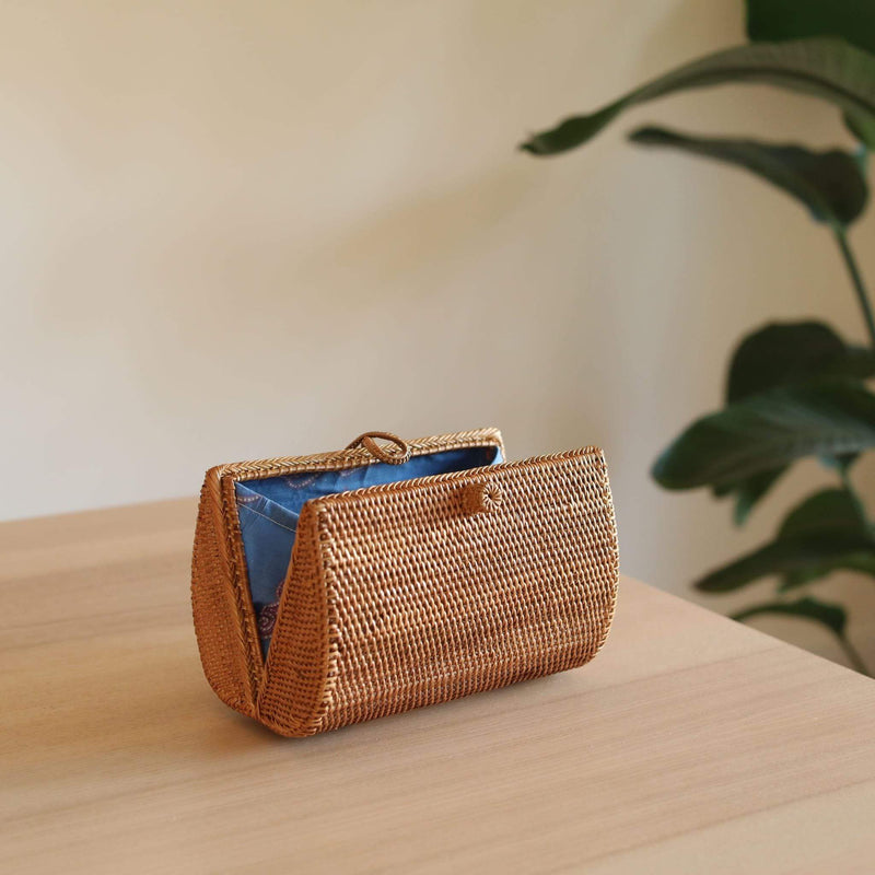 A beautiful, handmade rattan clutch by Ganapati Crafts Co., resting on a wooden surface with a plant in the background. The rectangular shape features intricate batik patterns on its exterior, while the interior has a plain lining. The clutch showcases the craftsmanship of Balinese artisans.