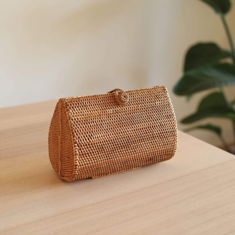 A woven rattan clutch rests on a wooden table amidst lush greenery, its natural material and eco-friendly design evident. The light brown clutch has a rectangular shape with a delicate weaving pattern, adorned with a unique batik lining. Made from Ata grass by skilled Balinese artisans in Bali, this handmade rattan bag features a hook closure and spacious interior, perfect for special occasions.