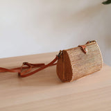 A woven rattan crossbody bag with a brown leather strap leans against a wooden surface, set against a simple yet stylish background featuring a white wall and a small plant. The bag's intricate rattan weaving is highlighted by soft natural lighting, showcasing its texture and bohemian charm.