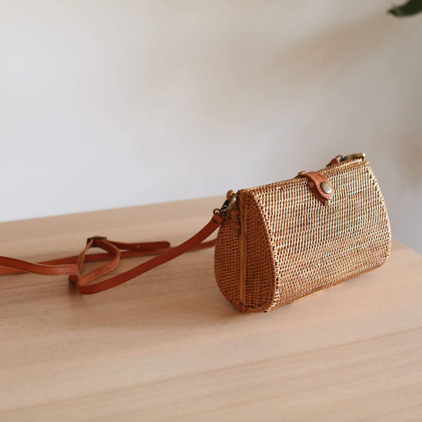 A woven rattan crossbody bag with a brown leather strap leans against a wooden surface, set against a simple yet stylish background featuring a white wall and a small plant. The bag's intricate rattan weaving is highlighted by soft natural lighting, showcasing its texture and bohemian charm.