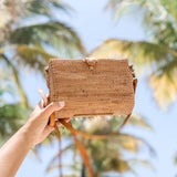 A person holds up a woven rattan crossbody bag, showcasing its textured surface and natural appeal. The bag is made from high-quality natural rattan Ata grass and features a detachable strap. Set against a bright blue sky with palm trees, the scene evokes a sunny beach or resort atmosphere.