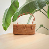 A woven rattan bag, made from natural rattan with a smooth finish, rests on a table or countertop. The light brown bag features a handle for carrying and a snap closure. A potted plant adds greenery nearby, suggesting versatility for daily use or formal events.