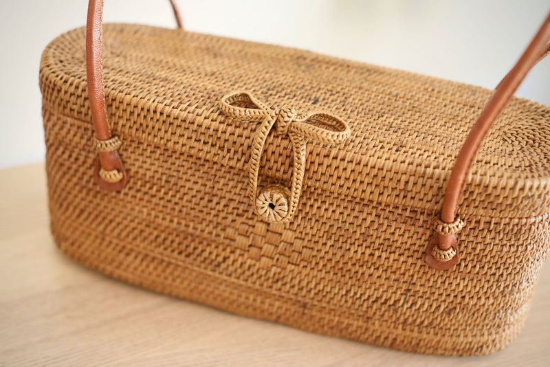 A woven, light brown wicker bag rests on a wooden surface, featuring a handle made from the same material as the body secured with a leather snap closure. A visible bow-tie detail adds decorative appeal. The bag's shape is petite and relaxed, with a smooth rattan finish that won't catch on clothing.