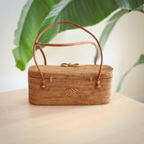 A woven, light brown handbag crafted from rattan material sits on a wooden surface, featuring a genuine leather handle. The bag's design and texture are prominently displayed, with a plant in the background adding a touch of greenery.