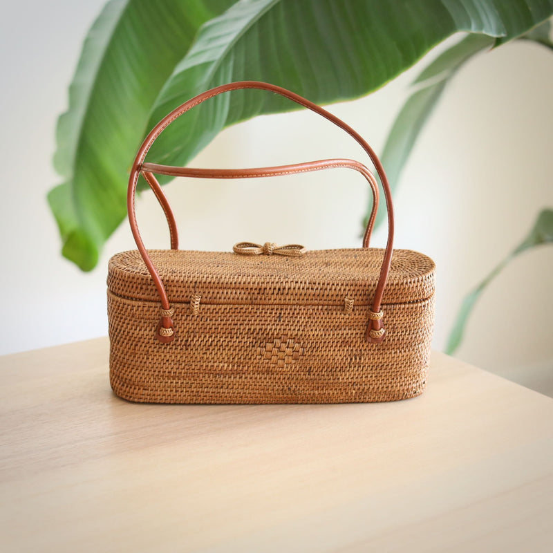 A woven, light brown handbag crafted from rattan material sits on a wooden surface, featuring a genuine leather handle. The bag's design and texture are prominently displayed, with a plant in the background adding a touch of greenery.
