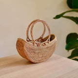 A woven basket with a round rattan handle rests on a surface, surrounded by an indoor setting with a plant in the background. The basket is made of natural rattan or a similar material, featuring a textured appearance due to its weaving pattern and earthy tone color. The product's main features include real rattan handles made from Ata grass, a smooth polished finish, and a unique Batik lining.