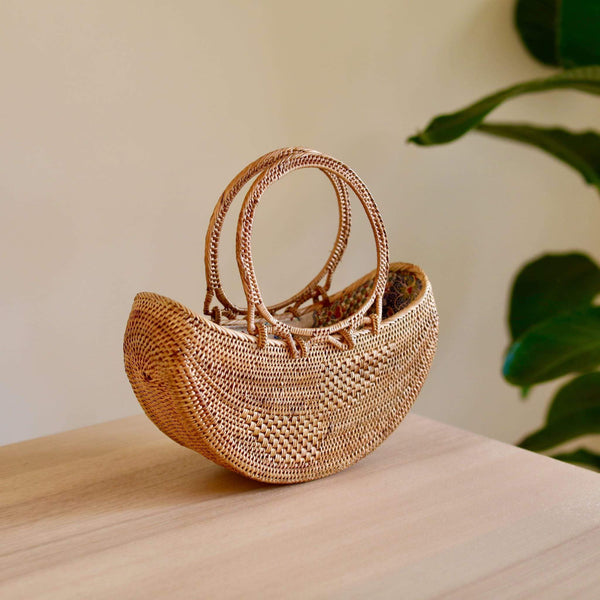 A woven basket with a round rattan handle rests on a surface, surrounded by an indoor setting with a plant in the background. The basket is made of natural rattan or a similar material, featuring a textured appearance due to its weaving pattern and earthy tone color. The product's main features include real rattan handles made from Ata grass, a smooth polished finish, and a unique Batik lining.