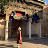 A woman wearing a hat carries a Bali Rattan Boat Clutch bag as she walks past a sign reading 'Race to Sun' in an outdoor setting with a traditional architectural structure featuring a wooden roof with intricate designs and blue fabric draping over the entrance. The bag, made from real rattan handles and Ata grass, has a smooth, polished finish and features a beautiful batik lining with a drawstring pouch.