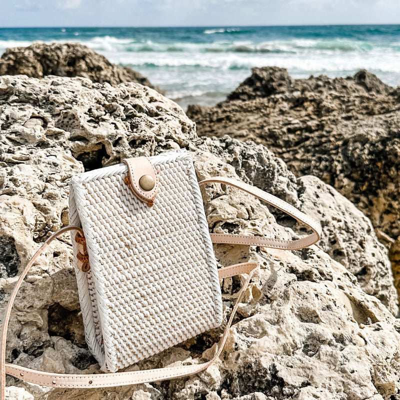 A beige woven rattan crossbody cellphone bag rests on a rocky surface near the ocean, surrounded by clear blue skies and calm waters. The bag features a high-quality woven rattan material with a natural texture, an adjustable strap made from real leather, and a secure snap closure. The lining is made of unique Batik cotton, adding to both style and durability.