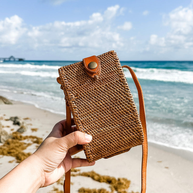 A person holding a woven rattan crossbody cell phone bag against a beach backdrop. The brown bag features a visible snap closure, indicating it's secure and adjustable, with a leather strap for comfort and versatility. The sandy beach background evokes a relaxed atmosphere.