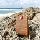 A woven rattan crossbody cellphone bag rests on a rocky surface near the ocean, surrounded by a clear sky and calm sea. The bag features a textured weave, metal clasp, and adjustable leather strap for secure carrying. Made from sustainable rattan with a smooth finish, this eco-friendly bag is perfect for travel and everyday use.