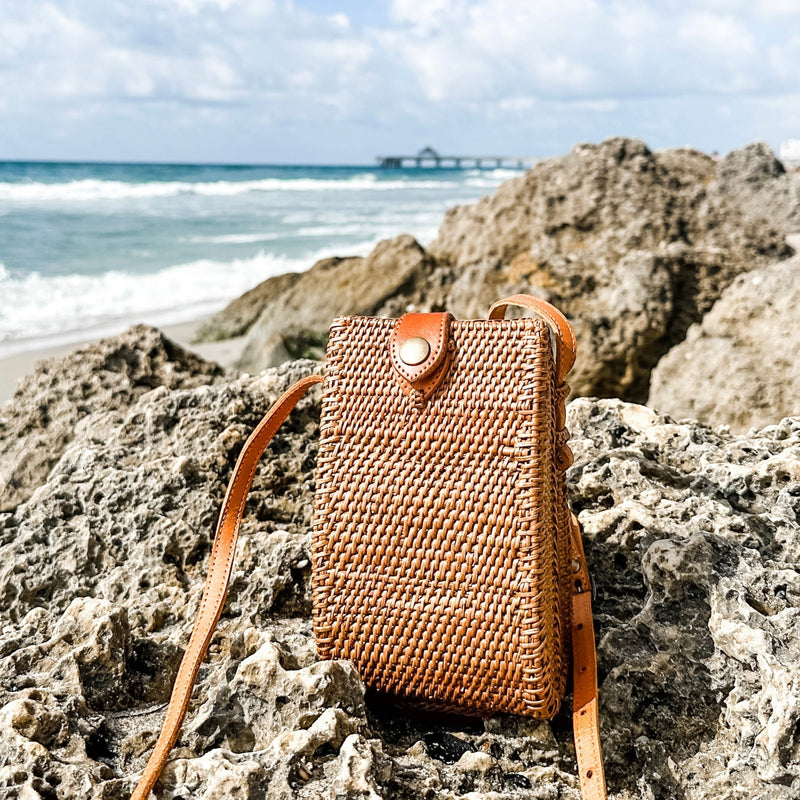 A woven rattan crossbody cellphone bag rests on a rocky surface near the ocean, surrounded by a clear sky and calm sea. The bag features a textured weave, metal clasp, and adjustable leather strap for secure carrying. Made from sustainable rattan with a smooth finish, this eco-friendly bag is perfect for travel and everyday use.