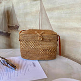 A woven rattan crossbody bag rests on a table, showcasing its light brown color and textured surface. The rectangular shape features rounded corners and an oval silhouette with intricate details. A secure leather adjustable strap allows for a perfect fit across the body. Made from high-quality rattan or bamboo, this stylish accessory is perfect for leisurely activities or everyday use.