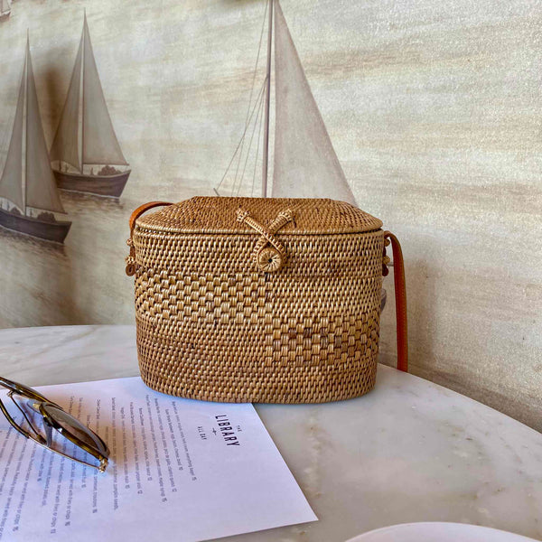 A woven rattan crossbody bag rests on a table, showcasing its light brown color and textured surface. The rectangular shape features rounded corners and an oval silhouette with intricate details. A secure leather adjustable strap allows for a perfect fit across the body. Made from high-quality rattan or bamboo, this stylish accessory is perfect for leisurely activities or everyday use.