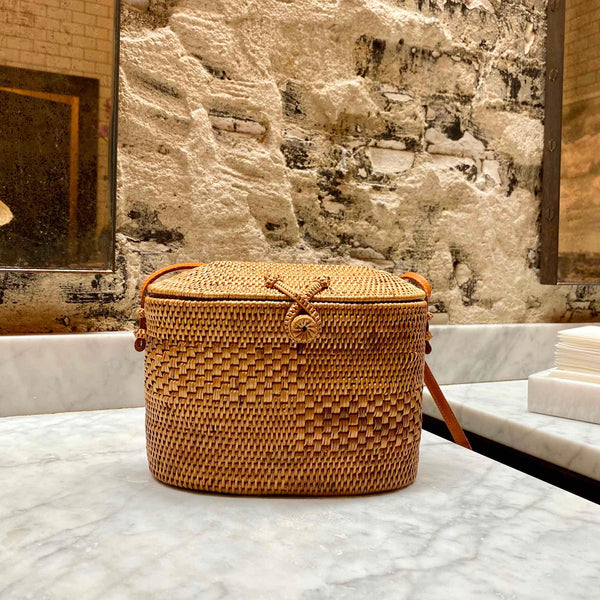 A light brown woven rattan crossbody bag rests on a surface, showcasing its elegant design and craftsmanship. The rectangular shape features rounded corners and a small decorative element on one corner, with a visible texture of the rattan material. A leather strap with a hook closure adds a touch of sophistication, while the background suggests an upscale indoor setting.