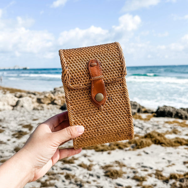 A model holds the Bali Rattan Crossbody Cellphone Bag - Ella, made of natural rattan material with a leather strap and snap closure. The bag features a smooth finish and is lined with Batik cotton for timeless style and durability.