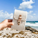A person holds up a woven rattan crossbody cellphone bag on a beach, showcasing its light-colored, textured surface and adjustable strap. The bag is made of natural materials like rattan and features a snap closure for secure phone carrying.