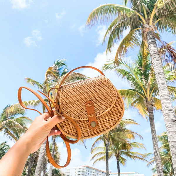 A handheld crossbody bag made from natural materials resembling Ata grass, adorned with a batik pattern resembling palm leaves. The bag features a real rattan strap that is adjustable and has a snap closure, resting on the arm of a person in the background who is holding it.