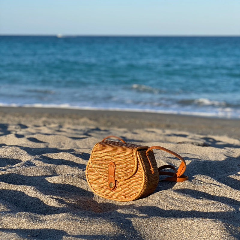 Bali Rattan Crossbody Bag - Guitar resting on sandy beach terrain, made from real Ata grass with a woven texture, featuring a natural rattan strap and Batik lining. The bag is placed in the foreground against a serene ocean backdrop, evoking a tropical vacation setting.