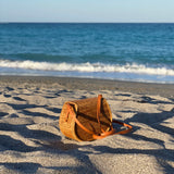 A woven Bali Rattan Crossbody Bag rests on a sandy beach, surrounded by the soothing backdrop of the ocean. The bag's natural brown and beige tones blend seamlessly with its surroundings. Its rattan strap is adjustable, adding a touch of rustic charm to any outfit.