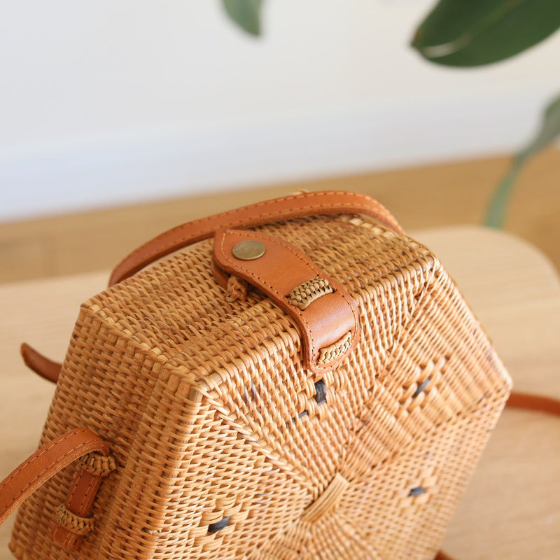 A woven rattan crossbody bag rests on a wooden surface, showcasing its hexagonal shape and natural materials. The bag features a brown leather strap with a metal buckle and a batik lining visible through the weave. This eco-friendly, handcrafted piece is made from Ata grass and has a unique design.