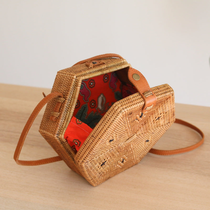 A woven rattan crossbody bag, hexagonal in shape, rests on a wooden surface, exuding a casual atmosphere. The bag features a real leather strap with an adjustable length and a unique batik lining. Made from natural rattan Ata grass, this eco-friendly handcrafted piece is perfect for everyday use.