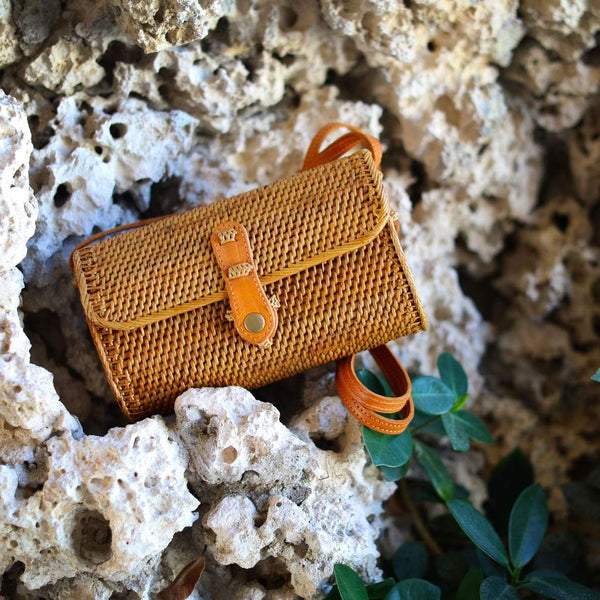 A small, woven rattan crossbody bag with a leather strap rests on a rocky surface amidst vegetation. The light brown bag has a rectangular shape with rounded corners, featuring a visible snap closure and adjustable leather strap that contrasts with the natural texture of the rattan material.