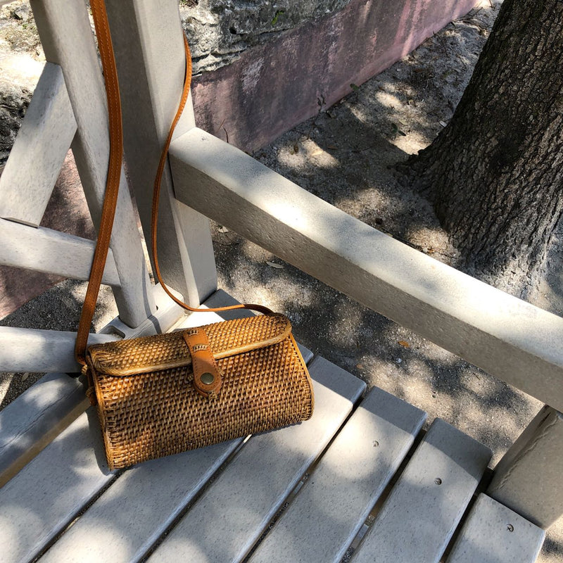 A small crossbody bag with rattan texture, placed on the armrest of a wooden bench. The bag features a brown leather strap that appears adjustable and a matching snap closure. The background suggests an outdoor setting.