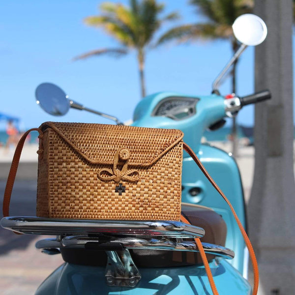 The Bali Rattan Crossbody Bag - Sophia rests on the seat of a parked motor scooter, surrounded by palm trees. The bag is made from handcrafted Ata grass with an adjustable leather strap for carrying. It features a compact design measuring 8.6