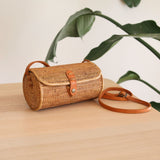 A woven crossbody bag sits on a wooden surface, its cylindrical shape tapering at the top. The light brown color suggests rattan or wicker, with darker brown lines and dots creating a textured pattern. A leather strap adds contrast to the design. In the background, a plant with broad leaves contributes to a natural atmosphere.