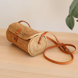A woven rattan crossbody bag rests on a wooden surface, its cylindrical shape and top handle visible. The adjustable leather strap and snap closure are also featured. A plant sits nearby on the white wall background.