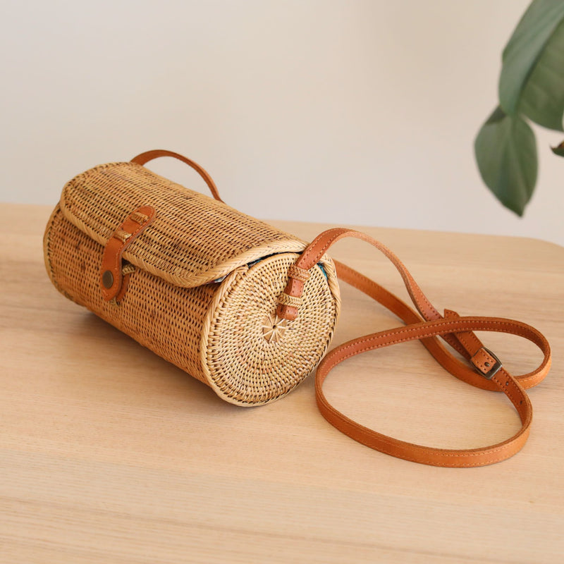 A woven rattan crossbody bag rests on a wooden surface, its cylindrical shape and top handle visible. The adjustable leather strap and snap closure are also featured. A plant sits nearby on the white wall background.