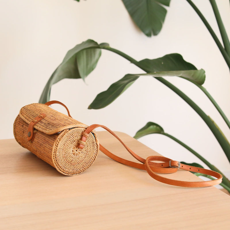 A woven Bali Rattan Crossbody Bag sits on a surface, surrounded by a plant's lush greenery. The bag itself features a natural rattan texture, eco-friendly material, and a cylindrical shape. A snap closure secures the bag, while an adjustable leather strap provides comfortable carrying. Inside, a beautiful batik lining adds elegance.