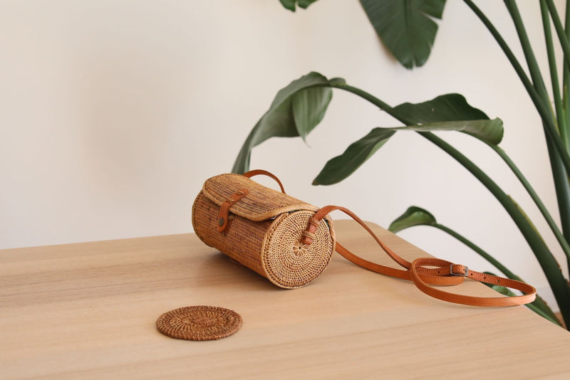 A Bali Rattan Crossbody Bag rests on a wooden surface, showcasing its cylindrical shape and textured rattan exterior. The bag features an adjustable real leather strap and a snap closure. A small circular object and a potted plant are nearby, adding to the relaxed atmosphere.