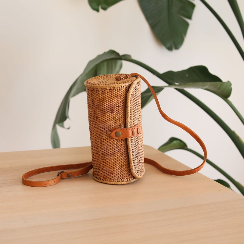 A woven crossbody bag, made from rattan or similar material, sits on a white surface. The cylindrical-shaped bag features a natural brown color with a bamboo leaf pattern, giving it a bohemian aesthetic. An adjustable leather strap and snap closure are visible on the front side, while a batik-lined interior is hinted at within.