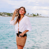 A woman stands on a serene beach, smiling at the camera while holding a woven straw bag made from natural materials. The bag features a simple design with a single strap over the shoulder.
