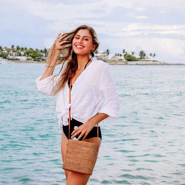 A woman stands on a serene beach, smiling at the camera while holding a woven straw bag made from natural materials. The bag features a simple design with a single strap over the shoulder.