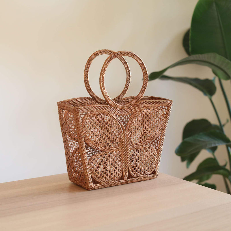 A wicker handbag featuring a butterfly pattern on its surface, resting on a wooden table amidst a serene backdrop of a plant and part of a white wall. The bag is made from rattan with two circular handles at the top, showcasing a trapezoid shape and unique butterfly design.