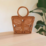 A woven rattan handbag with a butterfly pattern rests on a table, showcasing its trapezoidal shape and rattan handle. The background is minimalistic with a plant adding a touch of greenery. This eco-friendly bag features real rattan handles made from Ata grass, perfect for everyday use or a night out.